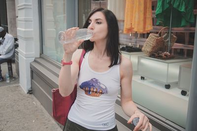 Woman drinking water while standing on footpath against store in city