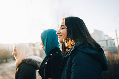 Side view of smiling teenage girl with young muslim friends in city against sky during sunset