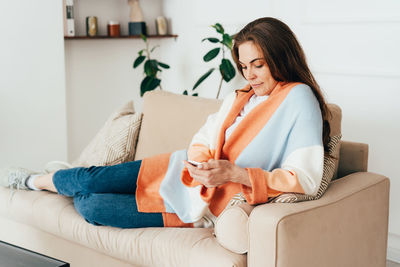 Young redhead woman sitting on the couch uses the phone to chat.